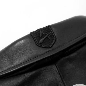 K&ouml;nnte beinhalten: Nahaufnahme einer schwarzen Lederjacke. Die Jacke hat einen kleinen, gestickten Aufn&auml;her mit einem stilisierten Emblem. Das Leder wirkt strukturiert, und die N&auml;hte sind entlang der N&auml;hte sichtbar. Die Gesamt&auml;sthetik ist schlicht und modern.