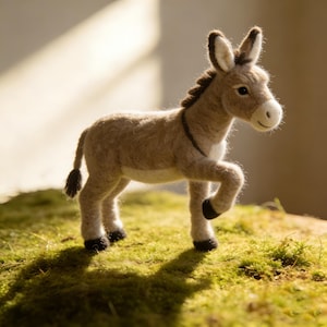 May include: A handcrafted felt donkey figurine in shades of tan, brown, and white. The donkey has a detailed mane, ears, and tail, and is posed walking on a bed of green moss. The figurine is approximately 6 inches long.