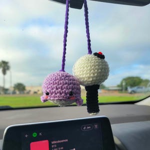 Puede incluir: Un adorno de coche tejido a ganchillo que presenta una ballena de color morado claro con ojos negros y mejillas rosadas junto a un bastón de luz esférico blanco y negro con un botón rojo, colgados de hilos de lana morados del retrovisor.
