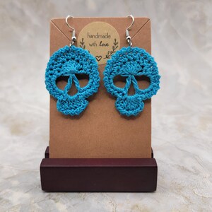 May include: A pair of turquoise crochet skull earrings with silver-colored hooks. The earrings are displayed on a brown card with a sticker that reads "handmade with love". The earrings are displayed on a dark brown wooden stand.