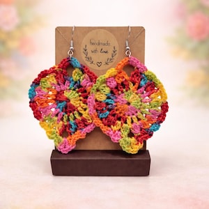 May include: A pair of handmade, colorful crochet earrings. The earrings are square-shaped with a floral pattern, featuring a vibrant mix of red, orange, yellow, green, and pink. The earrings are displayed on a brown stand with a tag that reads "handmade with love."