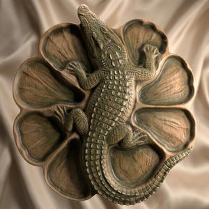 May include: A circular wooden serving tray featuring a carved crocodile relief in the center. The tray is divided into eight distinct, petal-shaped compartments surrounding the reptile, finished in a natural brown and muted green wood stain.