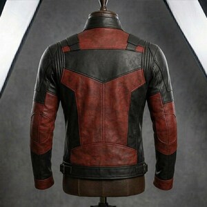 May include: A leather jacket with a two-tone design, featuring a combination of black and burgundy panels. The jacket has a classic biker style with a stand-up collar, shoulder accents, and a belt at the waist. The jacket is displayed on a mannequin.