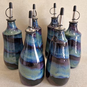 May include: A collection of five ceramic oil dispensers featuring a glossy glaze with horizontal bands of deep purple, navy blue, and sage green. Each bottle is topped with a stainless steel pour spout and a black plastic cap with a tether.