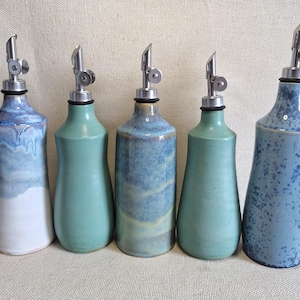May include: Five ceramic oil dispensers with stainless steel pour spouts. The bottles feature various blue and green glazes, including solid matte teal, blue and white drip patterns, and a speckled blue finish, all arranged in a single row.