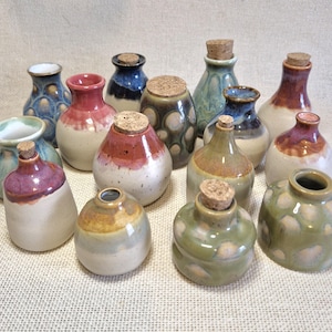 May include: A collection of fifteen small ceramic bottles in various shapes and sizes, featuring diverse glaze colours including blue, green, pink, and brown. Several bottles are topped with small, round, light brown cork stoppers.