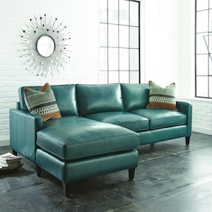 Könnte beinhalten: Ein teal-farbenes Ledersofa mit einer Chaiselongue. Das Sofa hat zwei dekorative Wurfkissen mit geometrischen Mustern. Der Raum verfügt über eine weiße Backsteinmauer, ein großes Fenster und einen dekorativen Sonnenstrahlspiegel.