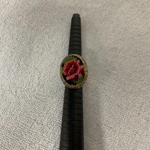 Könnte beinhalten: Ein ovaler Ring mit einem floralen Stickmotiv aus einer roten Rose und grünen Blättern, eingefasst in einen goldfarbenen Metallrahmen mit texturiertem Rand. Der Ring ist auf einem schwarzen, konischen und gerillten Ringstock platziert.