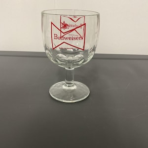 Pode incluir: Taça de vidro transparente com haste e taça larga. A taça tem um logótipo Budweiser vermelho e branco impresso na frente. O copo está sobre uma superfície cinza escuro.