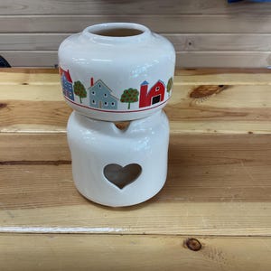 May include: A two-piece white ceramic oil warmer featuring a top bowl decorated with a colorful pattern of houses, barns, and trees. The base section is a solid white cylinder with a heart-shaped cutout on the front for ventilation and light.