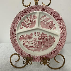 May include: A round, white ceramic divided plate featuring a red transferware pattern of a landscape with pagodas, trees, and birds. The rim is decorated with a geometric border pattern. The plate is displayed on a gold-toned metal stand.