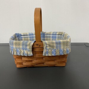 May include: A rectangular woven wooden basket featuring a single upright handle. The interior is lined with a blue, beige, and white plaid fabric that folds over the top rim, creating a pleated edge detail around the entire basket opening.