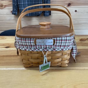 Può includere: Un cesto in legno intrecciato con finitura marrone chiaro, dotato di un coperchio in legno abbinato con pomello centrale. Il cesto è foderato con un tessuto a quadri rossi, verdi e bianchi e include un'etichetta in metallo argentato Longaberger 2004.