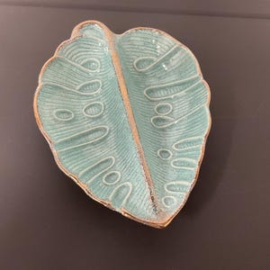 Op de afbeelding: Een bladvormige keramische schaal met een glanzend turquoise glazuur en een getextureerd oppervlak met verhoogde nerven en cirkelvormige patronen. De randen en de centrale rug van de schaal hebben een ruwe, ongeglazuurde bruine afwerking.