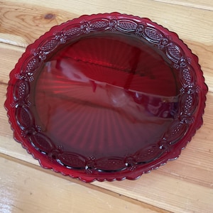 May include: A round, deep red glass plate featuring a scalloped edge and a decorative border with an embossed lattice and dot pattern. The centre of the plate has a smooth, flat surface with subtle radiating lines extending towards the rim.