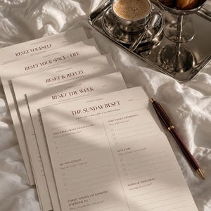 Könnte beinhalten: Ein Stapel cremefarbener Papierplaner mit schwarzem Text, darunter Titel wie The Sunday Reset, Reset The Week, Reset & Refuel, Reset Your Space + Life und Reset Yourself, begleitet von einem dunkelroten Stift, einem silbernen Tablett, Kaffee und Gebäck.