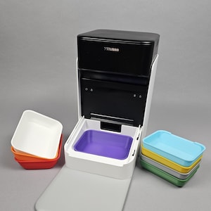 Può includere: Un distributore automatico di cibo per animali Petlibro bianco e nero con una vaschetta centrale viola. A sinistra ci sono ciotole rettangolari impilate bianche, arancioni e rosse. A destra ci sono ciotole azzurre, gialle, grigie e verdi.