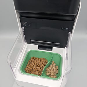 May include: A white and black Petlibro automatic pet feeder featuring a green, dual-compartment food tray. The left side of the tray contains dry brown kibble, while the right side holds wet food chunks. The device has a digital control panel.