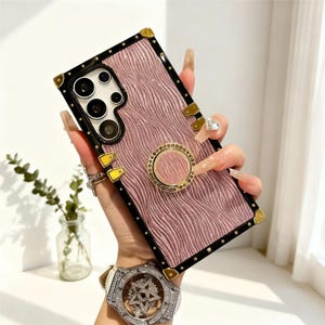 May include: Pink and black phone case with a textured design, gold accents, and a ring holder. The case has a black border with gold corner details and a gold ring holder. The phone case is designed to fit a smartphone.