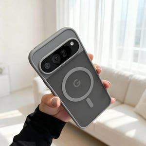 Puede incluir: Una funda gris para teléfono con un recorte para la cámara y un diseño circular con la letra "G". La funda tiene un acabado mate y un borde gris contrastante. La funda está diseñada para un teléfono inteligente.