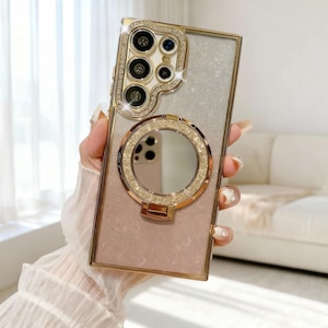 May include: A gold and glitter phone case with a ring holder and camera lens protector. The case has a gold frame and is decorated with rhinestones. The ring holder has a mirror. The case is designed for a smartphone.