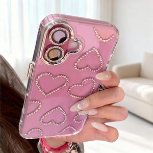 Puede incluir: Funda de teléfono rosa con borde transparente, adornada con brillantes pedrería en forma de corazón. La funda tiene un anillo de cámara plateado y una parte trasera transparente. Diseñada para un smartphone.