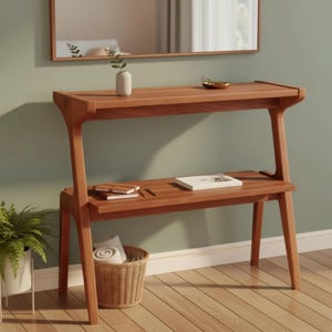 May include: A wooden console table with two shelves, a mirror, and a woven basket. The table is made of light brown wood and has a minimalist design. A small vase with a plant, a gold-colored bowl, notebooks, and a pen are on the shelves.