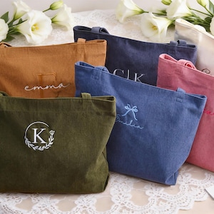 May include: Several corduroy tote bags in shades of brown, navy, pink, and olive green. Each bag has embroidered details, including names or initials. White flowers are arranged around the bags. Measurements are not available.