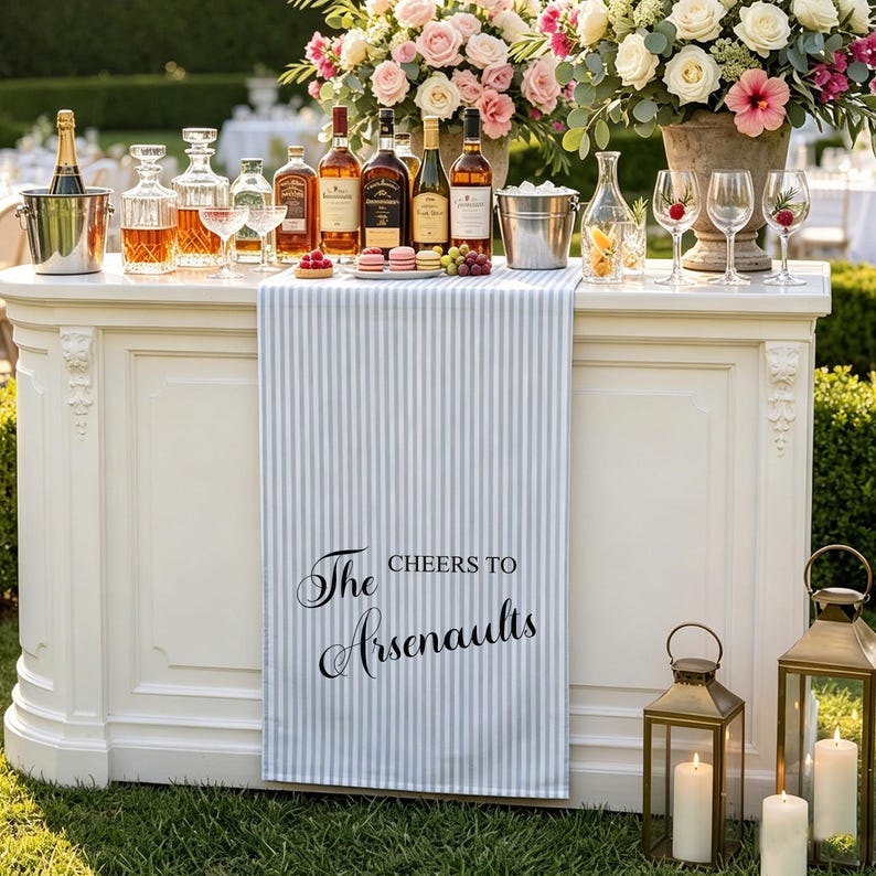 May include: A light blue and white vertical striped table runner featuring the black printed text 'Cheers to The Arsenaults' draped over a white bar. The bar is topped with various glass bottles, wine glasses, a metal ice bucket, and flowers.