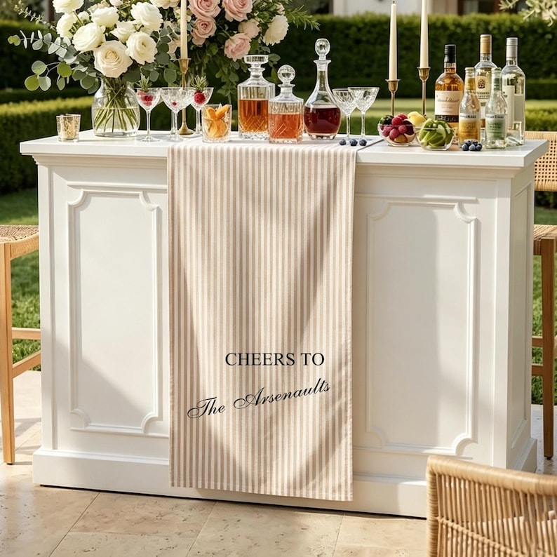 May include: A white bar table draped with a beige and white striped table runner featuring the black printed text CHEERS TO The Arsenaults. The table is topped with glass decanters, liquor bottles, crystal glassware, and a floral arrangement.