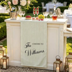 May include: A light green and white striped table runner featuring the black printed text The Williams and CHEERS TO. The runner is draped over a white bar counter decorated with a champagne bucket, wine glasses, fruit, and macarons.