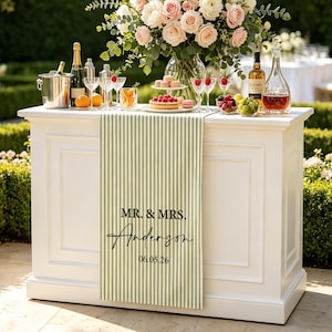 May include: A sage green and white vertical striped table runner draped over a white bar. The runner features black printed text that reads MR. & MRS. Anderson followed by the date 06.05.26 in a clean, centered font arrangement.