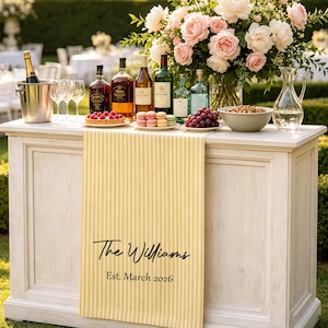May include: A yellow and white striped table runner featuring the black printed text The Williams and Est. March 2026. The runner is draped over a white wooden bar topped with wine bottles, a champagne bucket, glasses, fruit, and a floral arrangement.