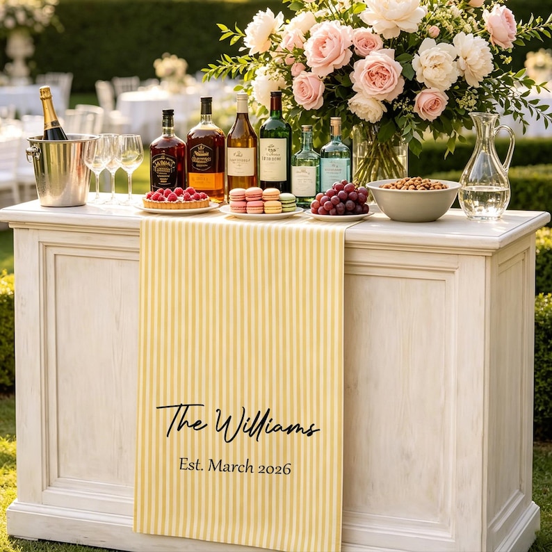 May include: A yellow and white striped table runner featuring the black printed text The Williams and Est. March 2026. The runner is draped over a white wooden bar topped with wine bottles, a champagne bucket, glasses, fruit, and a floral arrangement.