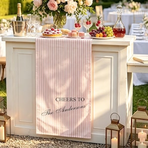 May include: A light pink and white vertical striped table runner featuring the black printed text CHEERS TO The Andersons. The runner is draped over a white wooden bar counter, which is decorated with a champagne bucket, fruit, and glasses.