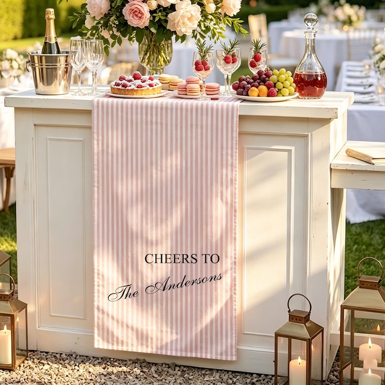 May include: A light pink and white vertical striped table runner featuring the black printed text CHEERS TO The Andersons. The runner is draped over a white wooden bar counter, which is decorated with a champagne bucket, fruit, and glasses.