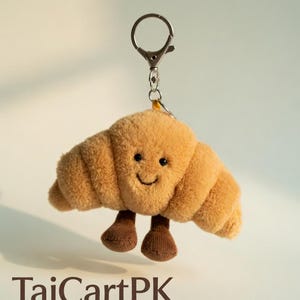 Pode incluir: Um chaveiro de croissant de pelúcia com um rosto sorridente e pequenos pés castanhos. O chaveiro tem um fecho e um anel prateados. O texto "TajCartPK" está na parte inferior da imagem.