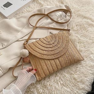 Könnte beinhalten: Eine gewebte Stroh-Clutch mit Klappenverschluss und einem langen, dünnen Schulterriemen. Die Tasche hat einen dekorativen Pompon und ist beige. Der Klappverschluss hat ein Halbkreismuster.