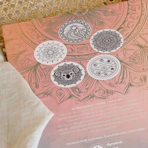 May include: A coloring book with a pink and beige mandala design. The cover features five circular illustrations of mandalas, including a koala, sun, and wave design. The text on the cover is in German.