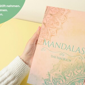 May include: A coloring book titled "Mandalas" is held by a person. The cover is a soft peach color with a mandala design in teal and gold. The text "Mandalas" is in teal script, and the words "Das Malbuch" are below.