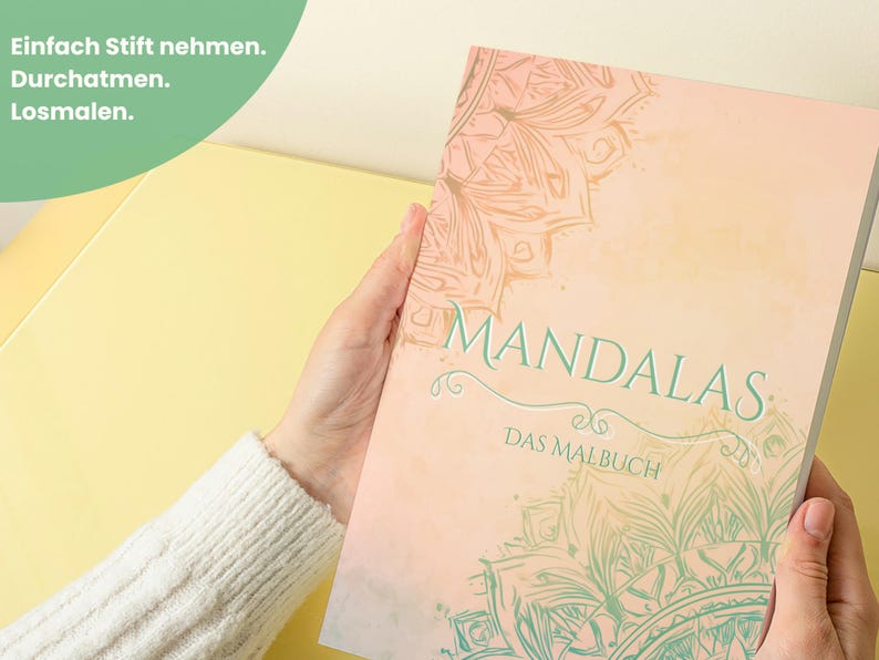 May include: A coloring book titled "Mandalas" is held by a person. The cover is a soft peach color with a mandala design in teal and gold. The text "Mandalas" is in teal script, and the words "Das Malbuch" are below.