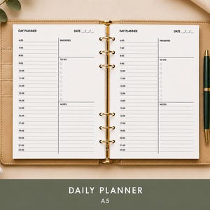 May include: Open daily planner with a tan leather cover and gold-colored rings. The planner is open to a blank page with a detailed schedule layout, including sections for priorities, to-do lists, and notes. A green pen rests on the right side.