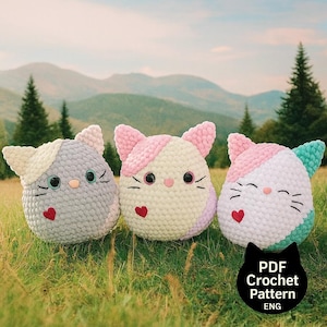 May include: Three handmade crochet cat pillows in a grassy field with mountains in the background. Each cat has a unique color scheme: gray, pink and white, and white with teal. They feature heart-shaped accents and embroidered faces. The image includes text: "PDF Crochet Pattern ENG."