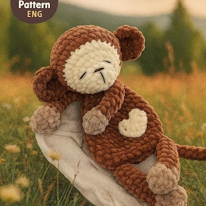 以下が含まれることがあります： クリーム色の顔と白いハート型のパッチが付いた、手作りの茶色の編み物モンキー。 モンキーは、緑の草と黄色の花が咲く野原の明るい灰色の表面に横たわっています。 画像には「PDF Crochet Pattern ENG」と書かれたロゴが含まれています。