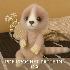 Puede incluir: Un juguete de suricato tejido a mano en tonos rosa y crema, sosteniendo una ramita de flores blancas. El suricato tiene grandes ojos negros y una nariz marrón. El texto "PDF CROCHET PATTERN ENG" está en la parte inferior.
