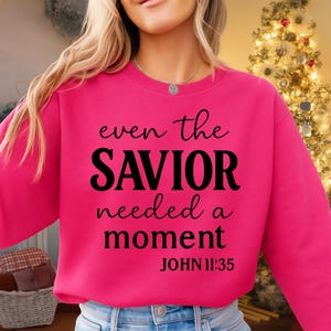 May include: A vibrant pink sweatshirt with the text "even the SAVIOR needed a moment" in black script. Below the text is "JOHN 11:35". The sweatshirt is a solid color and has a crew neck.