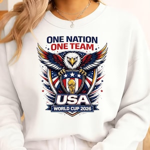 May include: White sweatshirt with a graphic of an eagle holding a shield with the text "USA" and "WORLD CUP 2026". The text "ONE NATION ONE TEAM" is above the eagle in blue and red.