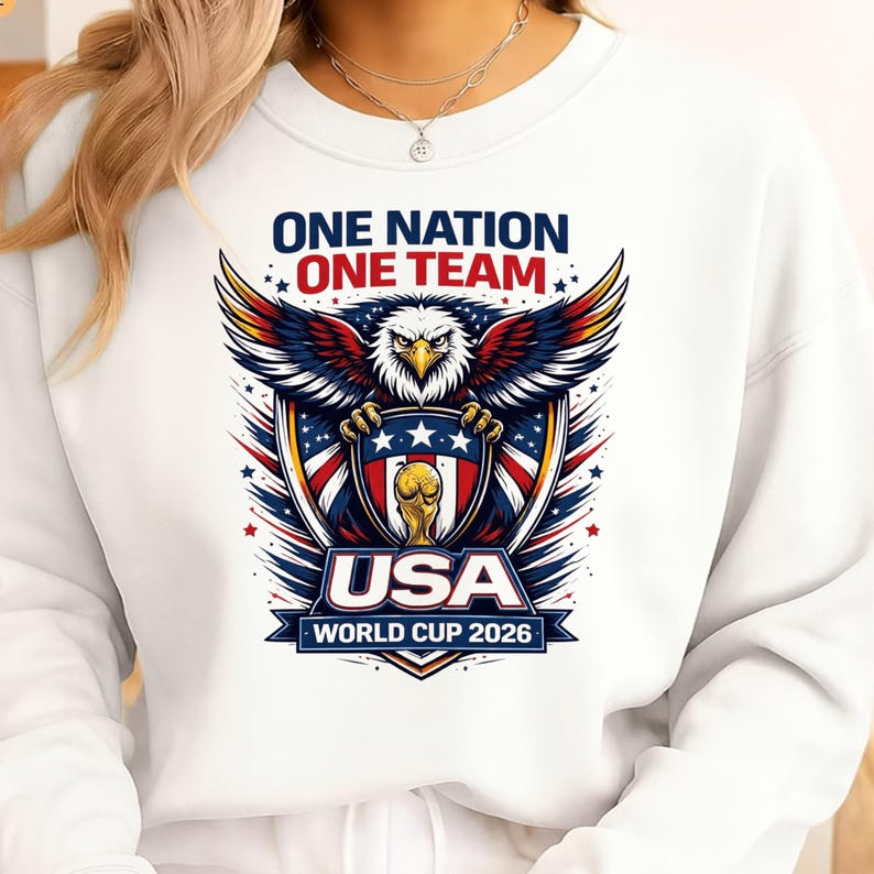 May include: White sweatshirt with a graphic of an eagle holding a shield with the text "USA" and "WORLD CUP 2026". The text "ONE NATION ONE TEAM" is above the eagle in blue and red.