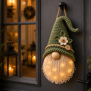 Peut inclure: Une décoration de gnome suspendue avec un chapeau en crochet vert olive texturé orné d'une fleur blanche et d'un bouton en bois. Le gnome possède un nez rond lisse et une longue barbe à franges claire illuminée par des LED blanc chaud.