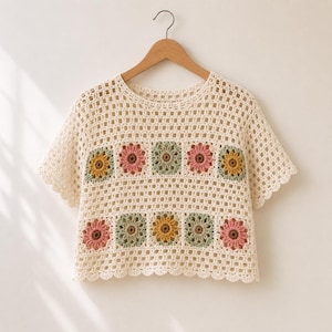 May include: A cream-colored crochet top featuring a grid-like openwork pattern and two horizontal rows of square floral motifs. The flowers are rendered in shades of dusty pink, sage green, and mustard yellow, with scalloped edges on the sleeves.